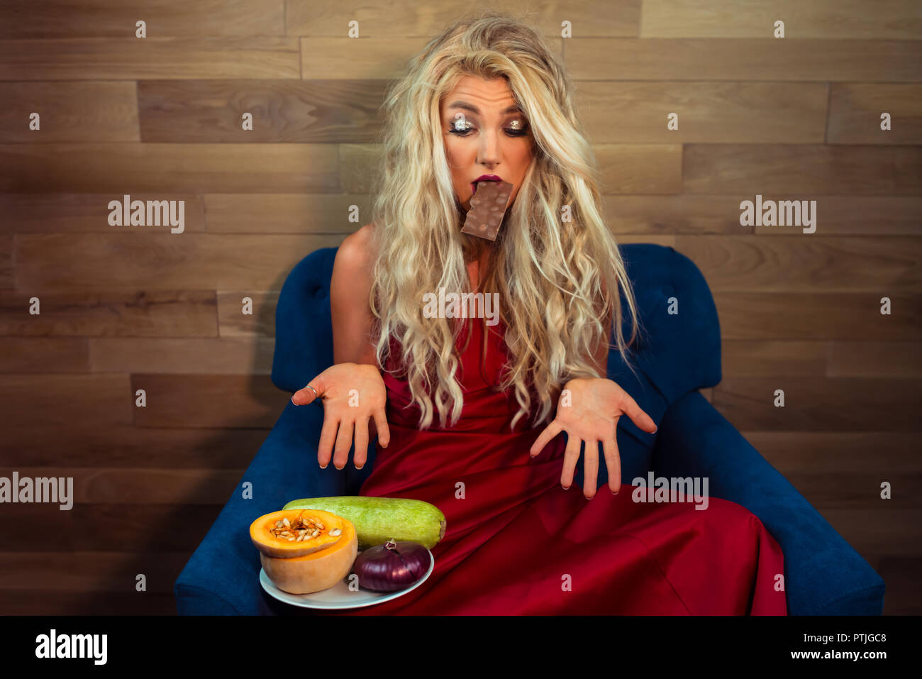 Junge Frau im eleganten roten Kleid sitzt auf einem Sessel mit Schokolade in den Zähnen und Suchen auf Gemüse. Lustige Hausfrau gestikulierend und Auswahl Stockfoto