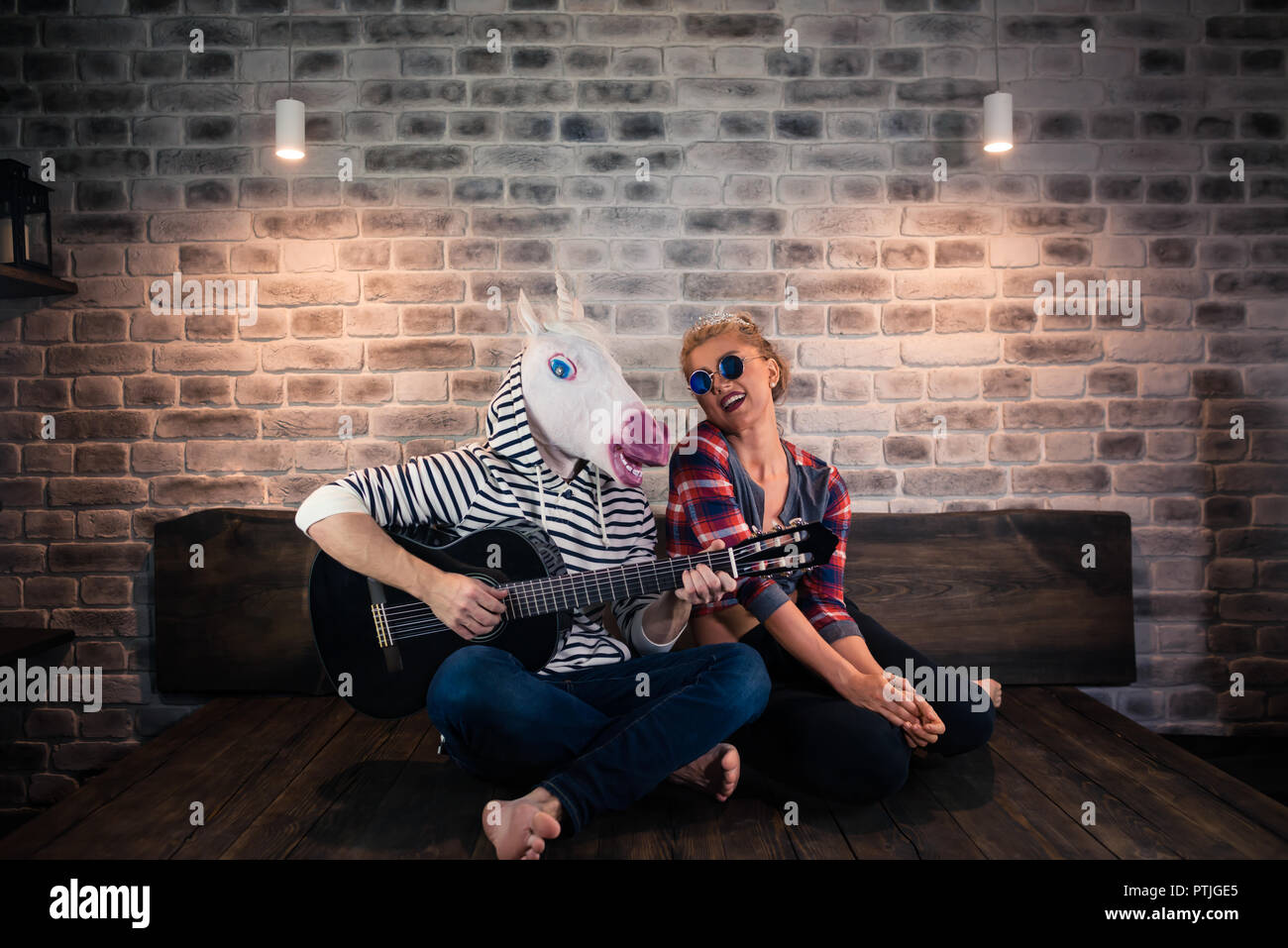 Freaky Mann in komischen Maske und kausalen Kleidung Wiedergeben von Musik auf Gitarre für schöne russische blonde Mädchen. Ungewöhnliche Paar zusammen in einem stilvollen Zimmer. Stockfoto