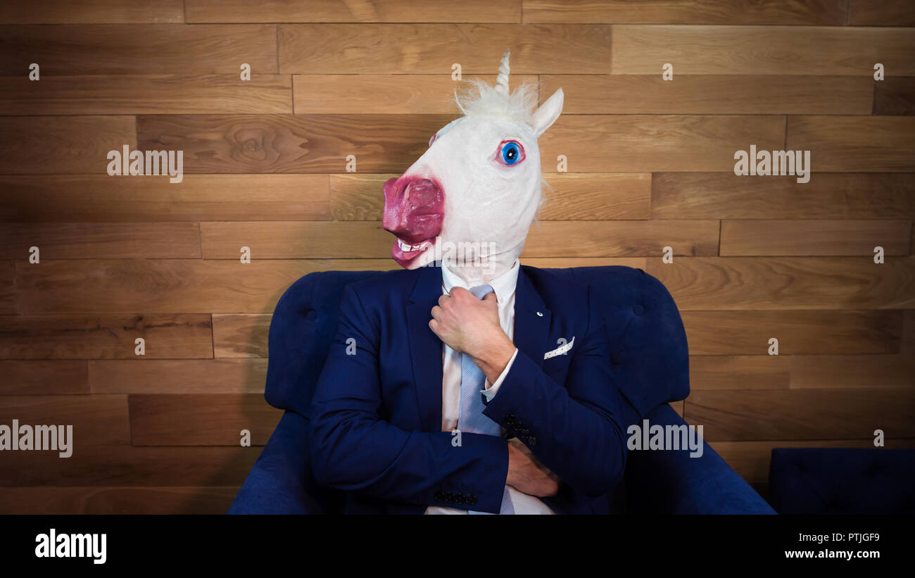 Portrait von ungewöhnlichen Einhorn im Home Office. Freaky junge Manager in komischen Maske auf dem Hintergrund der hölzernen Wand. Ernster Mann in Anzug sitzt wie Boss Stockfoto