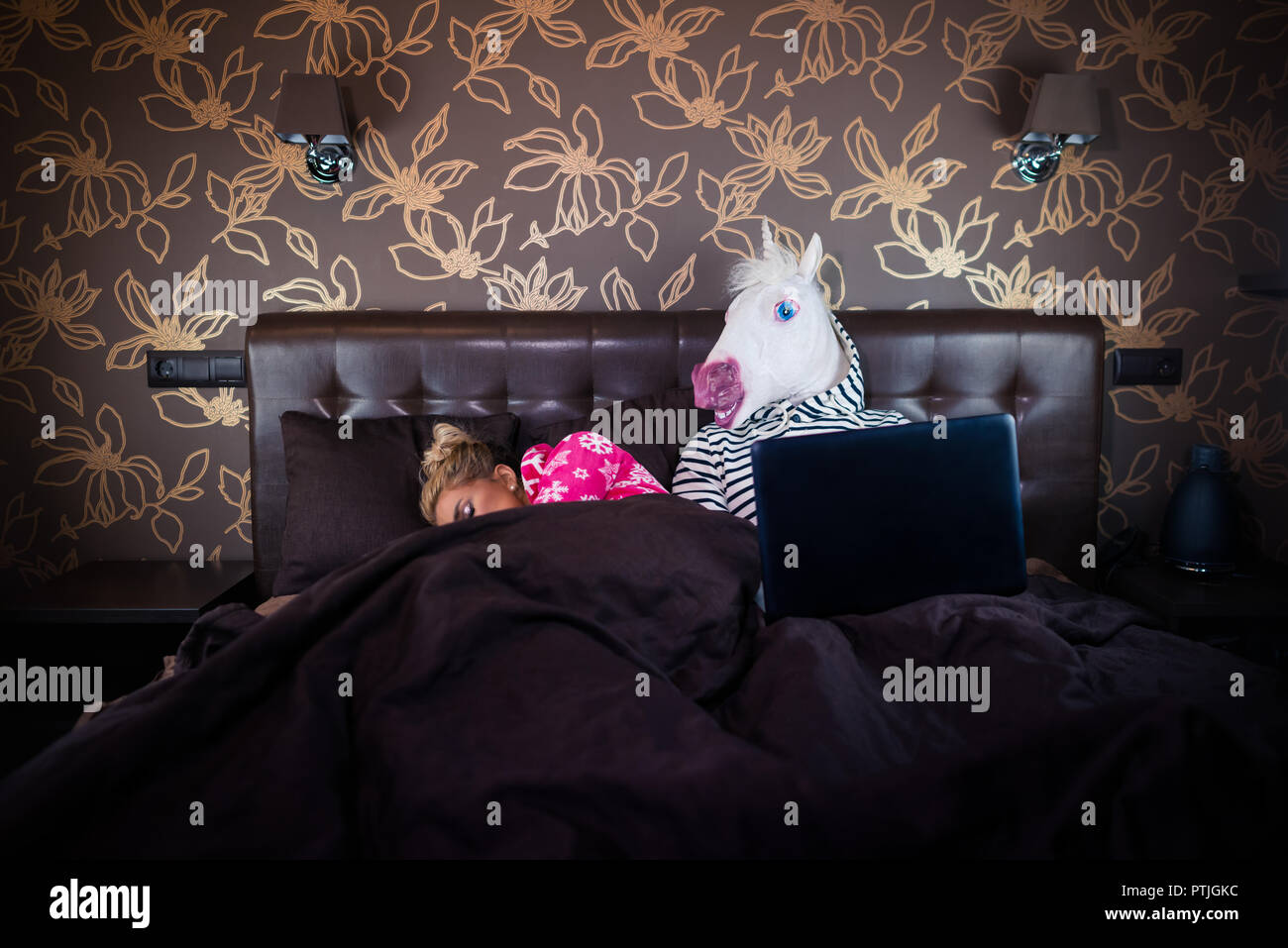 Blonde Mädchen schlafen mit lustigen Mann in komischen Maske. Ungewöhnliche Paar auf dem Bett liegend. Einhorn Film aufpassend auf Laptop in der stilvollen Schlafzimmer. Stockfoto