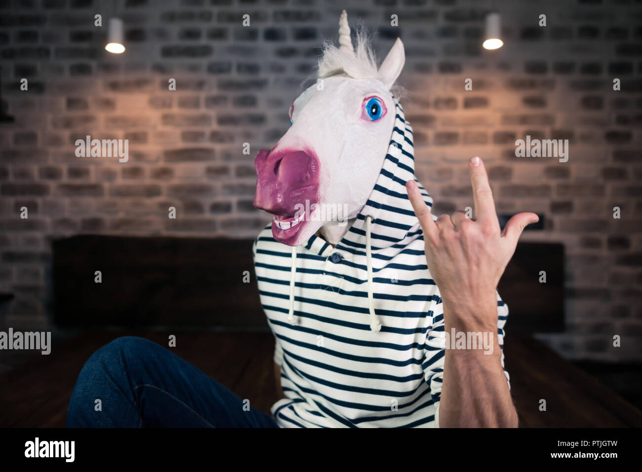 Freaky junger Mann in komischen Maske sitzt auf dem Bett in stilvolle Apartment. Lustige Einhorn in legere Kleidung zeigt Geste rock. Portrait von ungewöhnlichen Menschen. Stockfoto