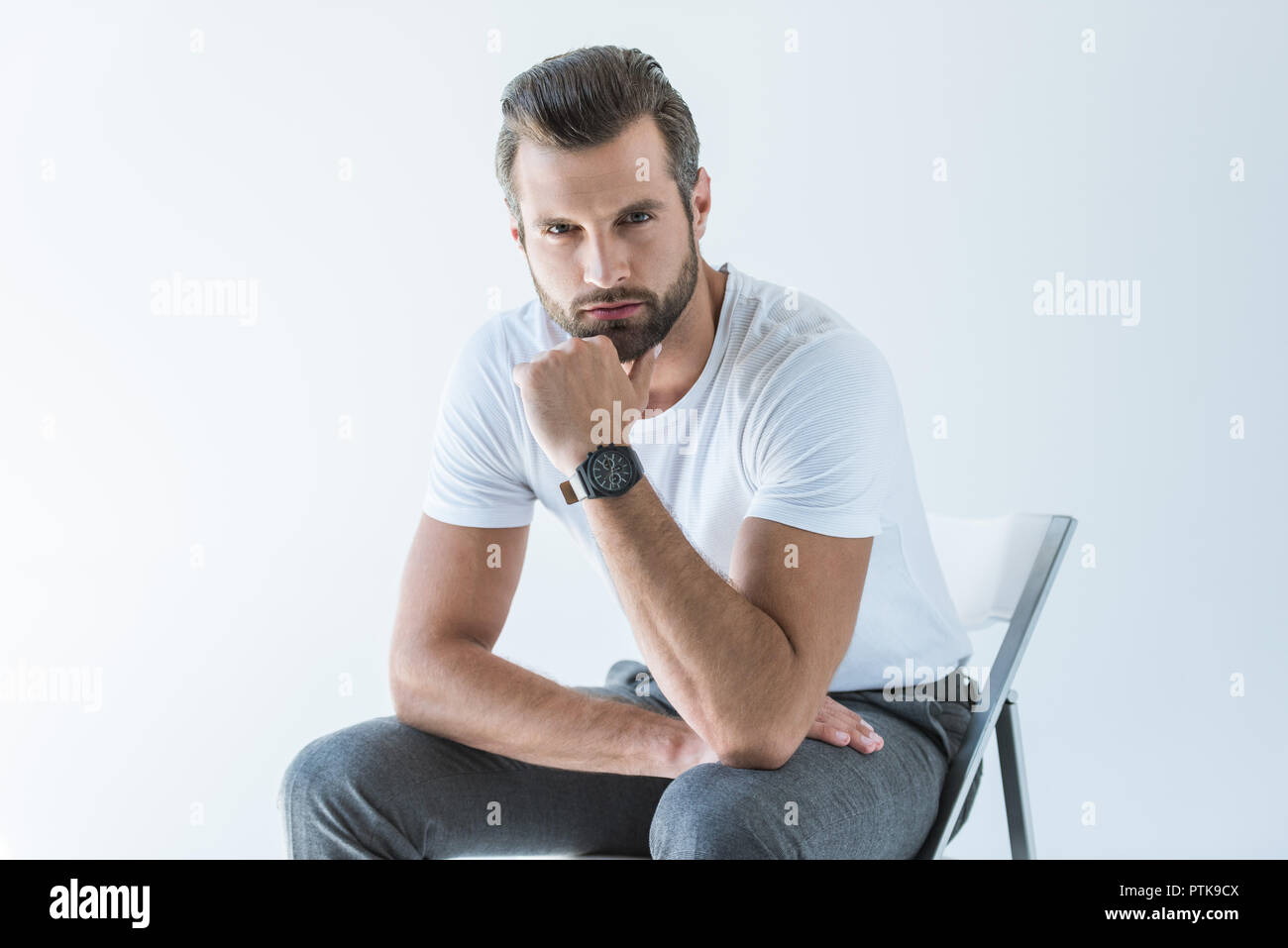 Modische gutaussehenden Mann im weißen T-Shirt mit Armbanduhr sitzt auf einem Stuhl, isoliert auf weißem Stockfoto
