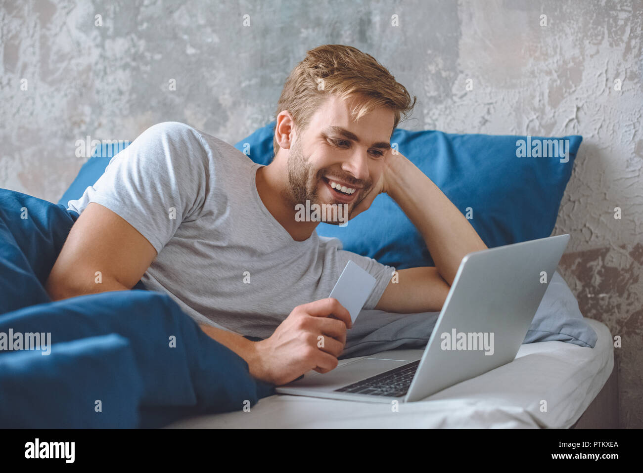 Lächelnden jungen Mann im Bett mit Kreditkarte, online shopping auf Laptop Stockfoto