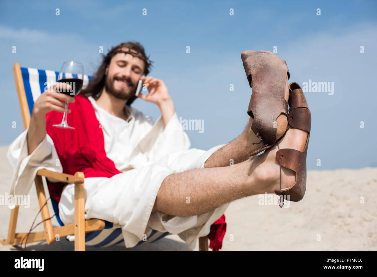 Lächelnd Jesus ruht auf einem Liegestuhl mit einem Glas Wein und sprechen durch Smartphone in der Wüste Stockfoto