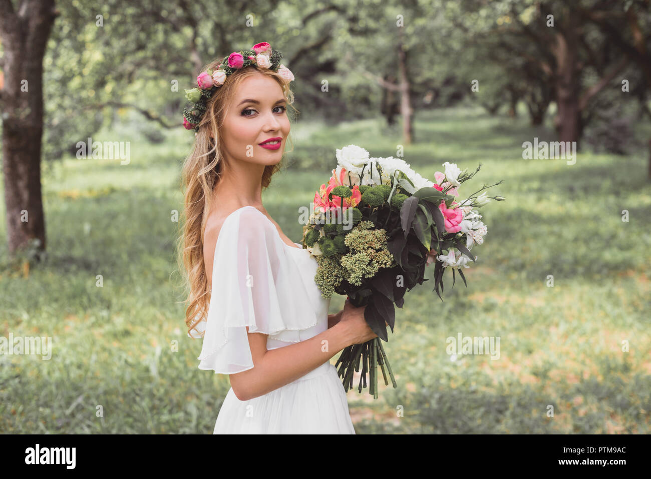 Glückliche Braut im Brautkleid und floralen Kranz holding Blumenstrauß und ein Lächeln auf die Kamera im Freien Stockfoto