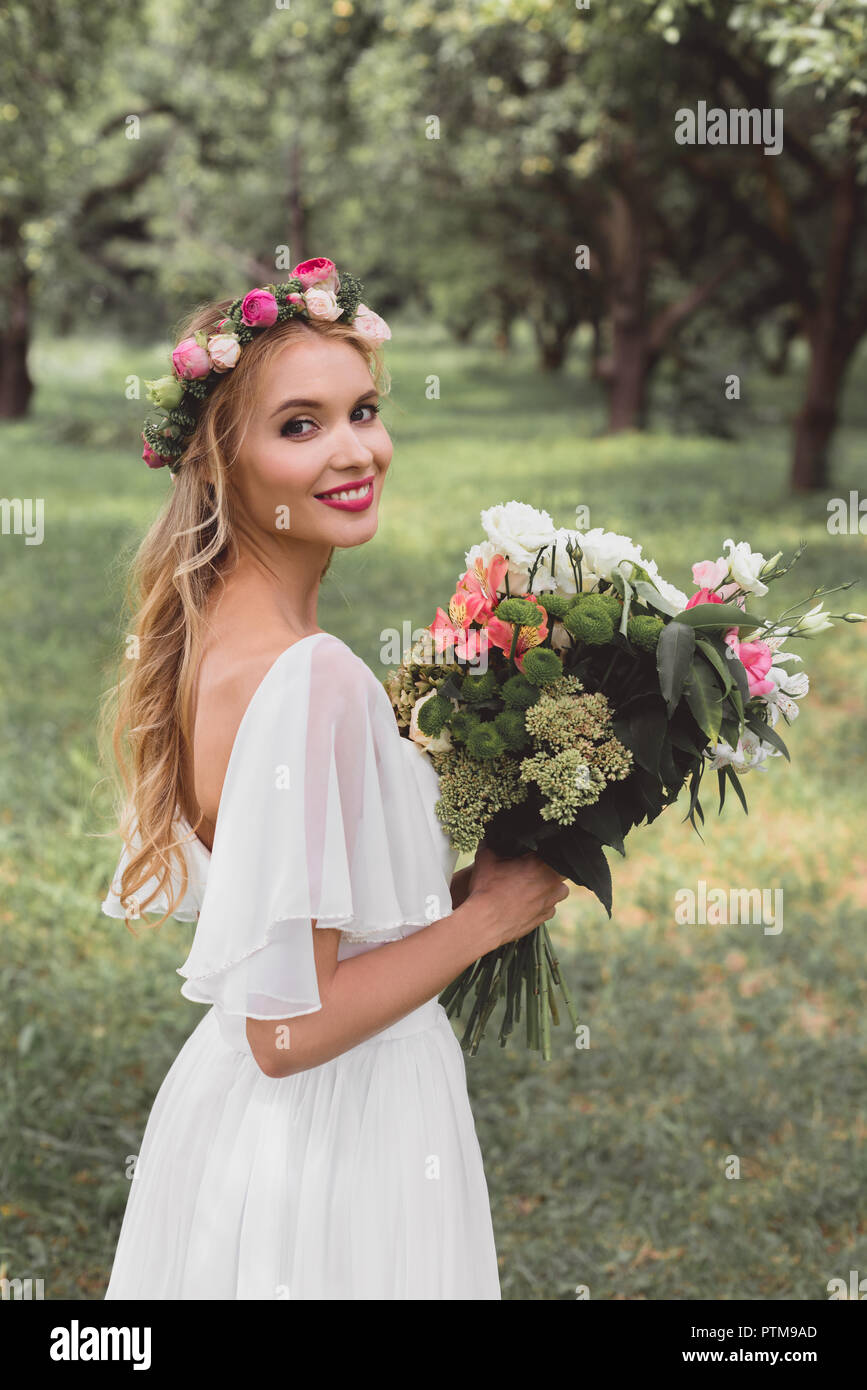 Gerne blonde Braut im Brautkleid und floralen Kranz holding Blumenstrauß und ein Lächeln auf die Kamera im Freien Stockfoto