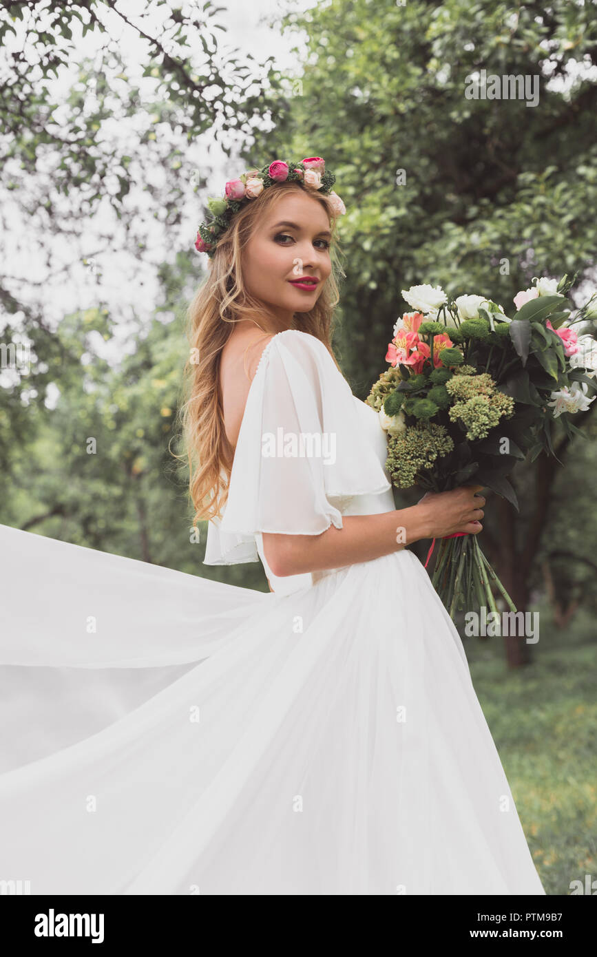 Schönen blonden Braut im Brautkleid und floralen Kranz holding Blumenstrauß und ein Lächeln auf die Kamera im Freien Stockfoto