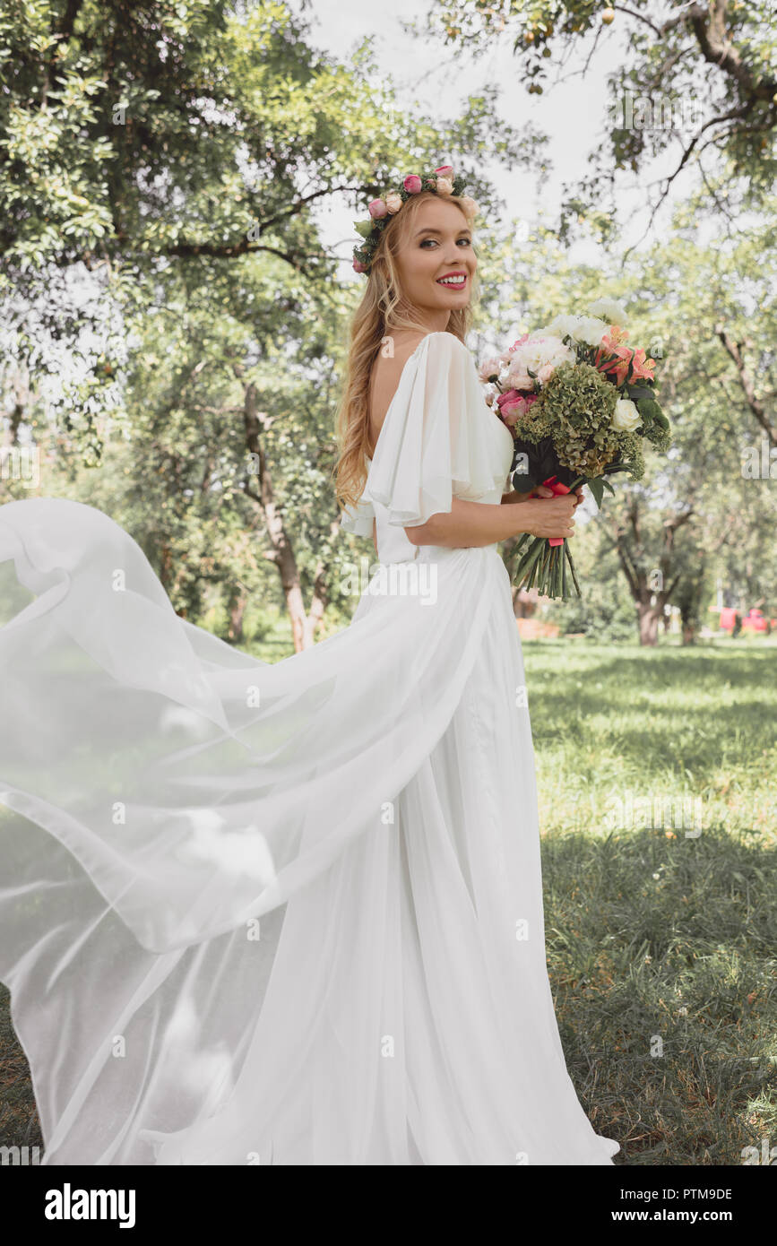 Schöne junge Braut im Brautkleid holding Blumenstrauß und ein Lächeln auf die Kamera im Park Stockfoto