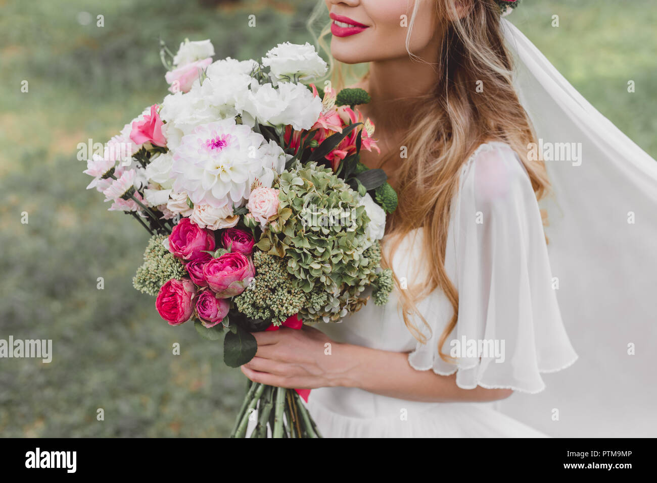 7/8 Schuß von lächelnden jungen Braut im Brautkleid und Schleier holding Blumenstrauß im Freien Stockfoto