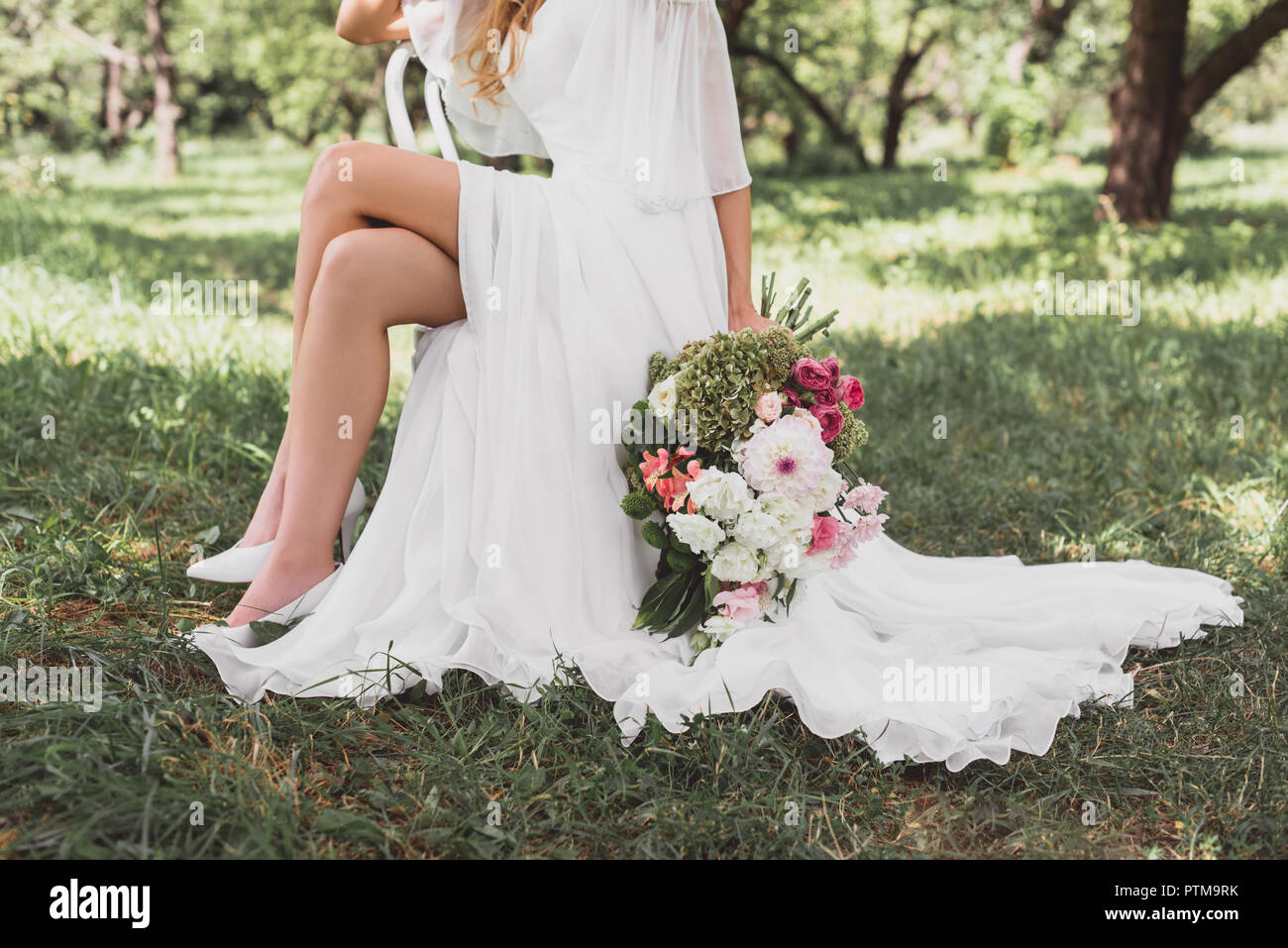 7/8 shot der Braut im Brautkleid sitzen auf einem Stuhl und halten Blumenstrauß im Freien Stockfoto