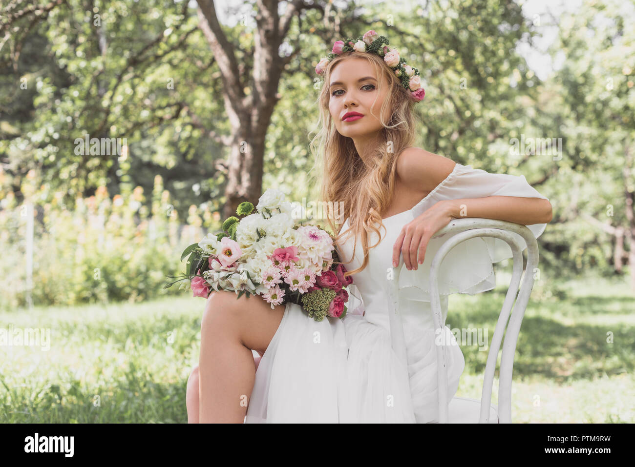 Schöne blonde junge Braut mit Hochzeit Kleid sitzt auf einem Stuhl und mit Blick auf die Kamera im Freien Stockfoto