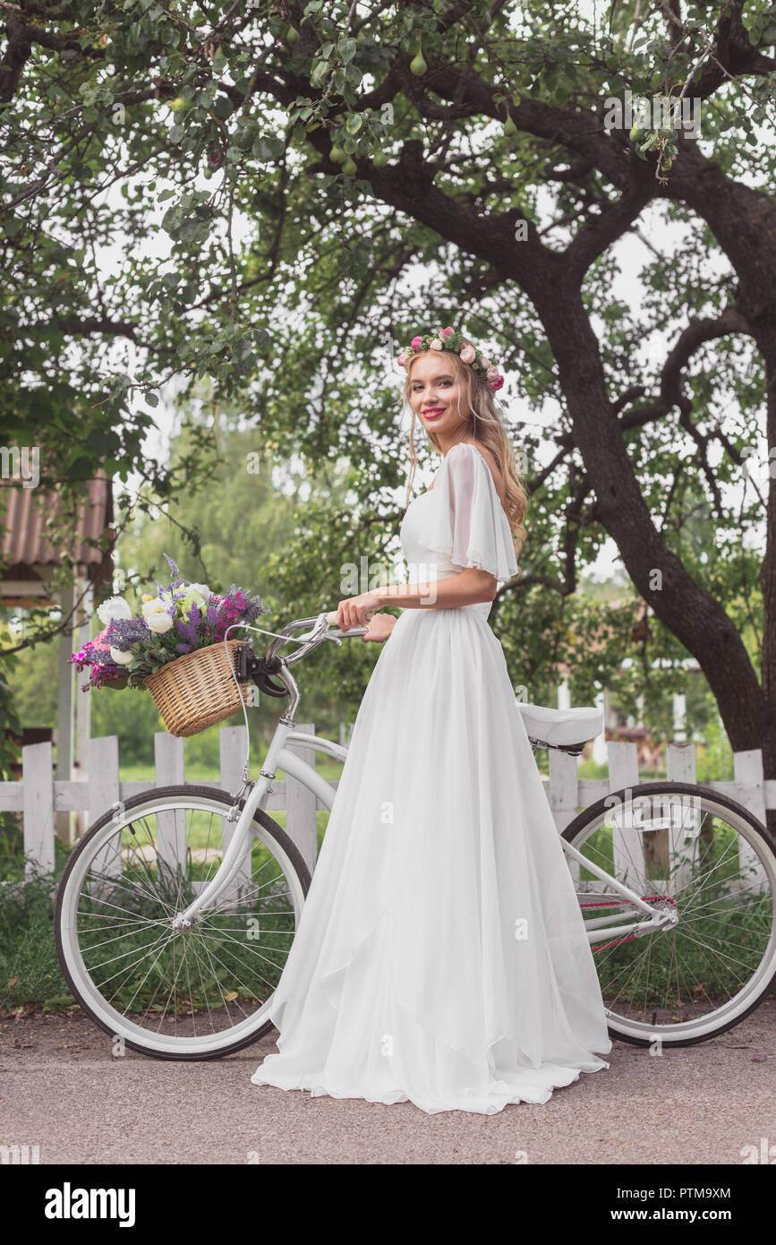 Schöne Braut in Blumen Kranz und Hochzeit stehend mit Fahrrad und lächelnd an Kamera Kleid Stockfoto