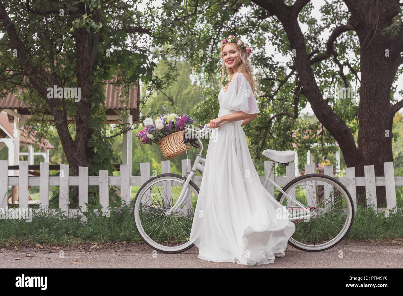 Junge Braut in Blumen Kranz und Hochzeit stehend mit Fahrrad und lächelnd an Kamera Kleid Stockfoto
