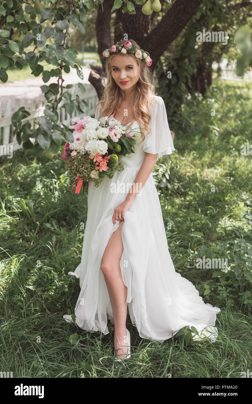 Schönen blonden Braut im Brautkleid und floralen Kranz holding Blumenstrauß und lächelnd an Kamera Stockfoto
