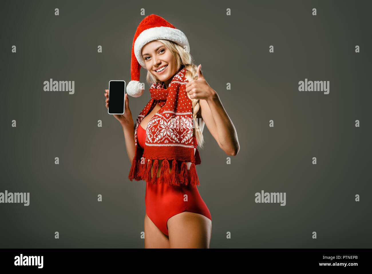 Lächelnde Frau in Rot swimming Suit, Schal und Santa Claus hat angezeigt Smartphone und Daumen hoch auf grauem Hintergrund Stockfoto
