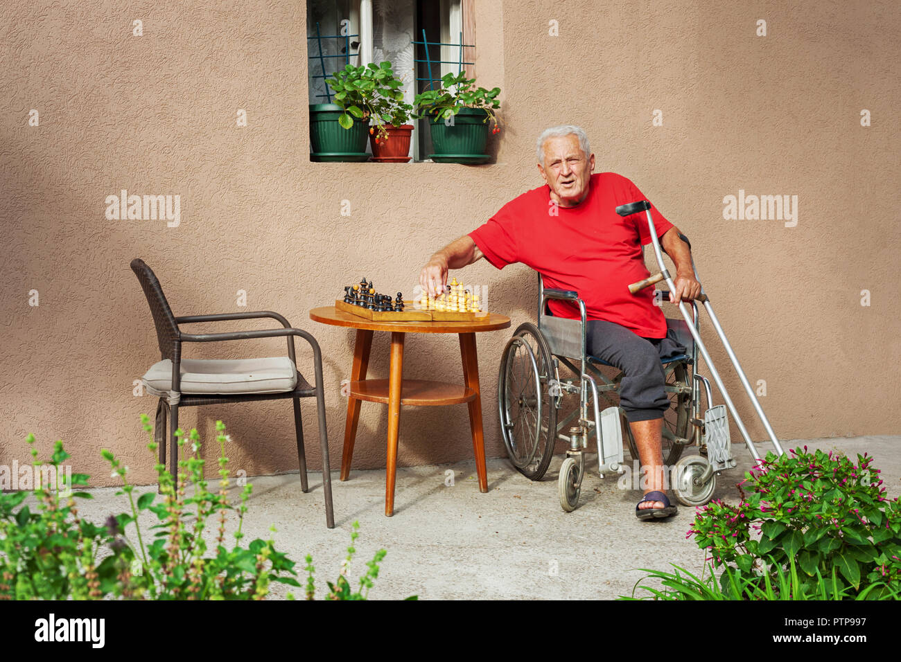 Älterer Erwachsener in einem Rollstuhl, mit Krücken, neben einem Schachbrett, an der Kamera, im Freien suchen, quadratischen Format Stockfoto