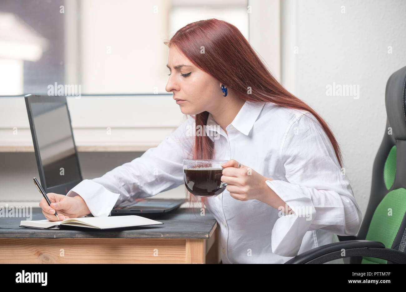 Eine junge, attraktive , Frau sitzt an einem Schreibtisch am Fenster. Sie arbeitet. Eine offene Laptop, Kaffee und Notebook sind auf dem Schreibtisch Stockfoto