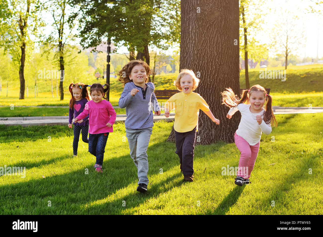 Eine Gruppe von kleinen glückliche Kinder laufen durch den Park, die im Hintergrund von Gras und Bäumen. Kinder Spiele im Freien, Ferien, Wochenende, Kinder- Da Stockfoto
