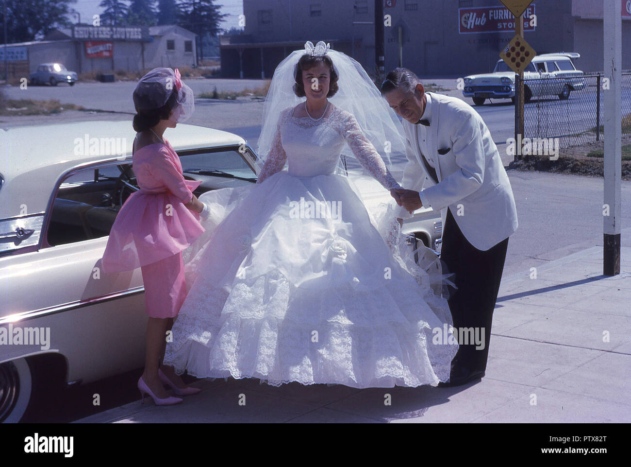 1962, historische, Kalifornien, USA, Sonnenschein, Hochzeit, junge Braut  strahlend in ihrem weißen Kleid kommt für Ihren großen Tag mit ihrer  Brautjungfer in Rosa und Vater in eine weisse Jacke ihr Begleiter  Stockfotografie -
