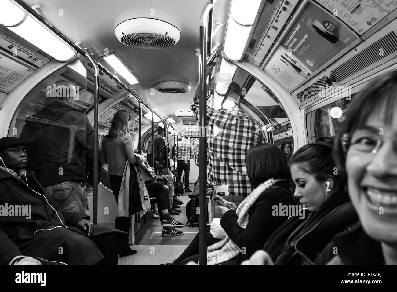 Schwarz & Weiß schoss der Fahrgäste in London das Rohr innerhalb des langen Waggon. Pendler Stehen und Sitzen ohne Interaktion; Spot der Tourist, der lächelt. Stockfoto