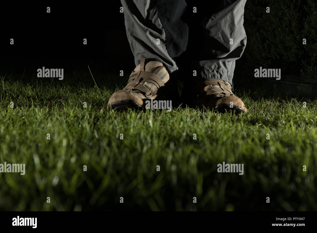 Niedrigen Winkel, Night Shot des Mannes Fuß in der Ausbilder, beleuchtet, stehend auf Gras im Garten. Der froschperspektive der männlichen Schuhe & Unterschenkel Hose in Dunkel. Stockfoto