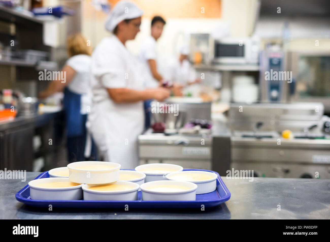 Viele Schüsseln mit Crème Brûlée und Mitarbeiter arbeiten in der Küche als Hintergrund Stockfoto