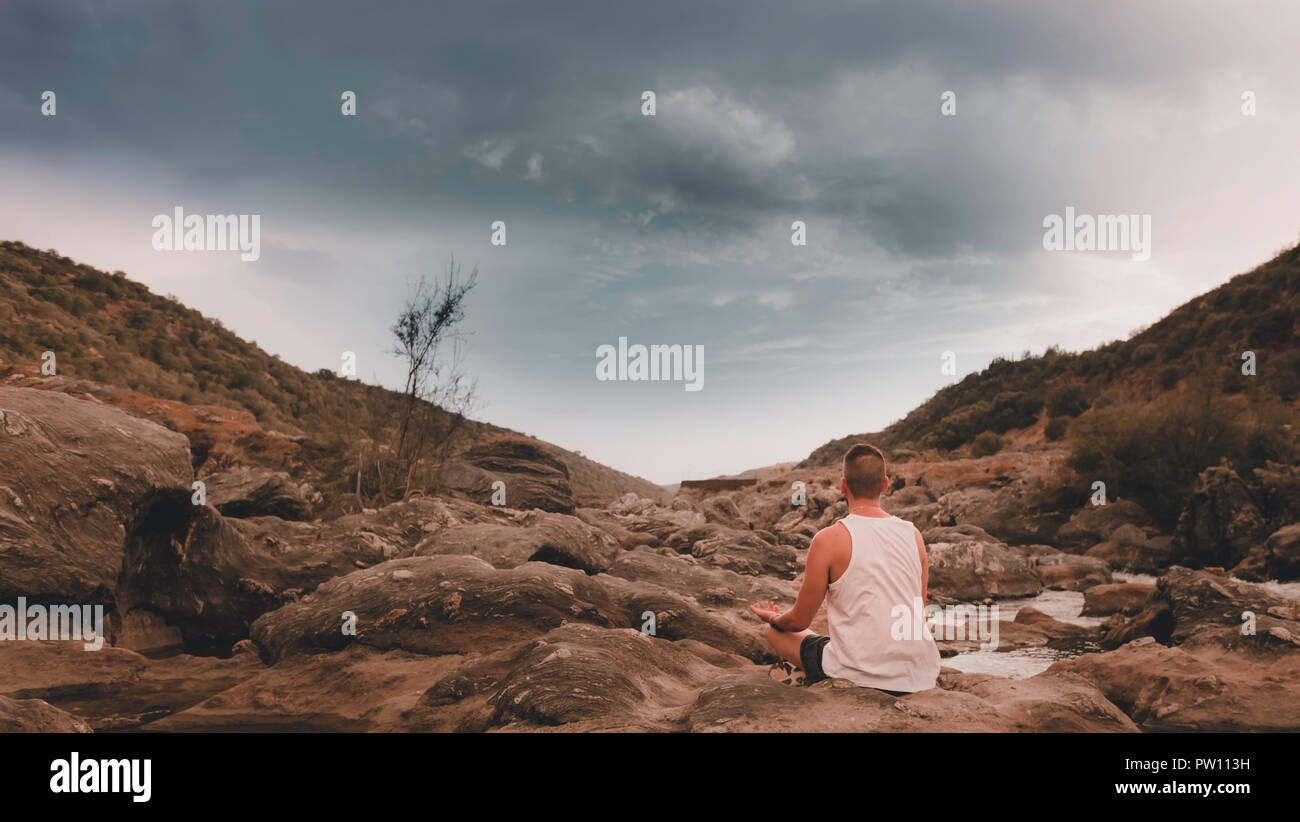 Junger Mann von hinten mit weißen Hemd Yoga auf dem Felsen in den Fluss Guadiana Stockfoto