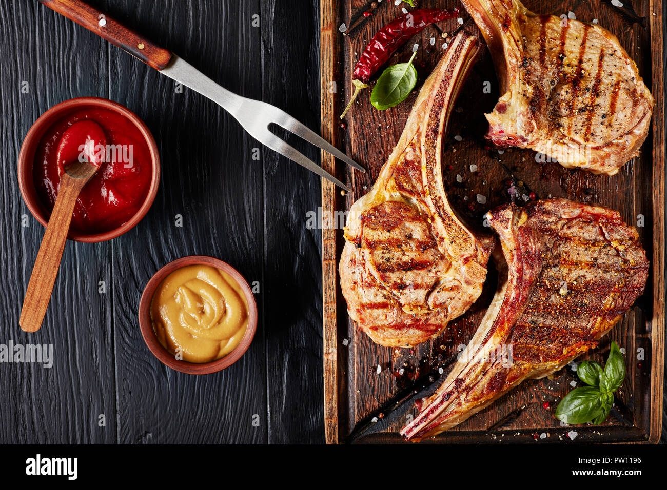 Drei trockenen Jahren tomahawk Steaks vom Grill oder Cowboy Rindersteaks auf einem alten unfreundlich Holz Schneidebrett mit Gabel auf einem Schwarzen Tisch mit Ketchup und Senf, Stockfoto