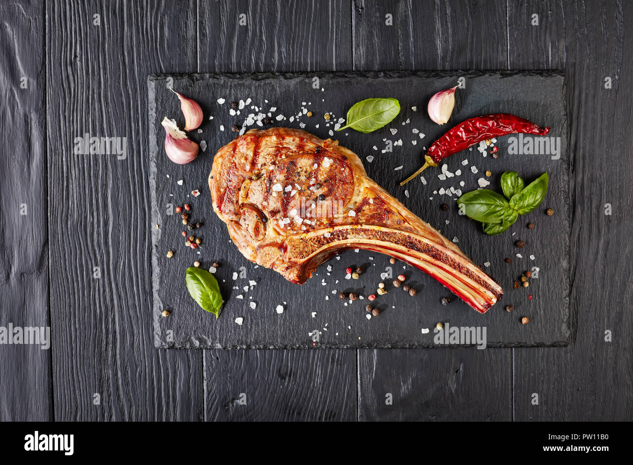 Close-up von gegrilltem Tomahawk Steak oder Cowboy Rindersteak auf schwarzem Schiefer mit Knoblauch, frischem Basilikum, Ansicht von oben, flach Stockfoto