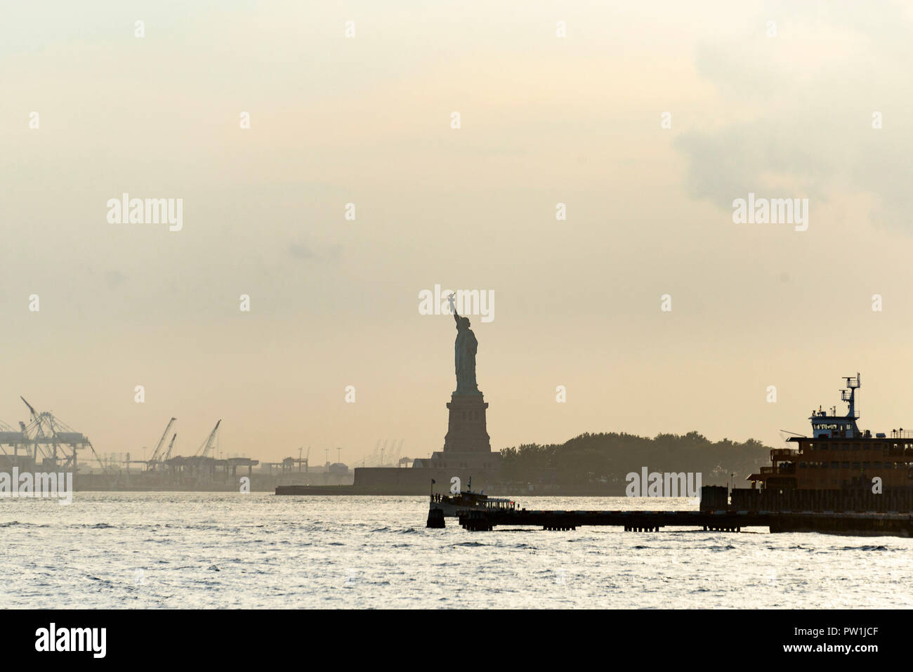 10-2018 Manhattan, New York. Die Freiheitsstatue in der Dämmerung, mit der Staten Island Ferry an der rechten, und Kräne und Docks von New Jersey auf der linken Seite. Foto: Stockfoto