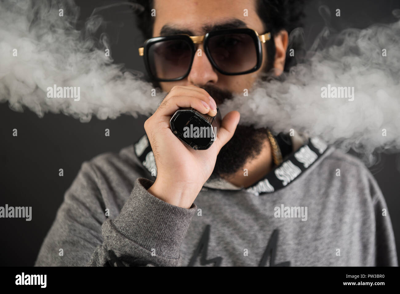 Portrait einer jungen Kerl mit Bart und Sonnenbrille und vaping eine elektronische Zigarette auf einem schwarzen Hintergrund. Stockfoto