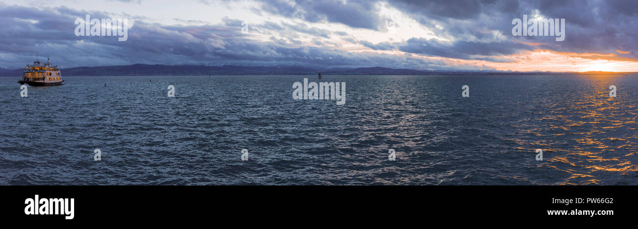 Panorama mit Sonnenuntergang über dem See Wasser und einzelne Boot segeln mit Passagieren, auf dem Bodensee in der Stadt Friedrichshafen, Deutschland Stockfoto