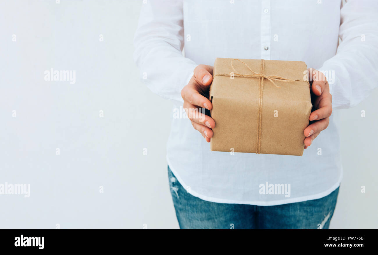 Junge kaukasier Frau in Jeans Shirt in den Händen hält, Geschenkbox in braun Handwerk Papier mit Garn gebunden. Weiße Wand. Weihnachten Neue Jahre Corporate p Stockfoto