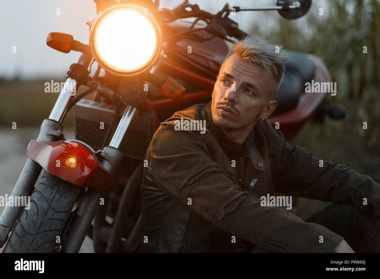 Brutaler Mann in einem militärischen Jacke sitzt und ruhen in der Nähe ein Motorrad mit Licht in das Feld am Abend Stockfoto