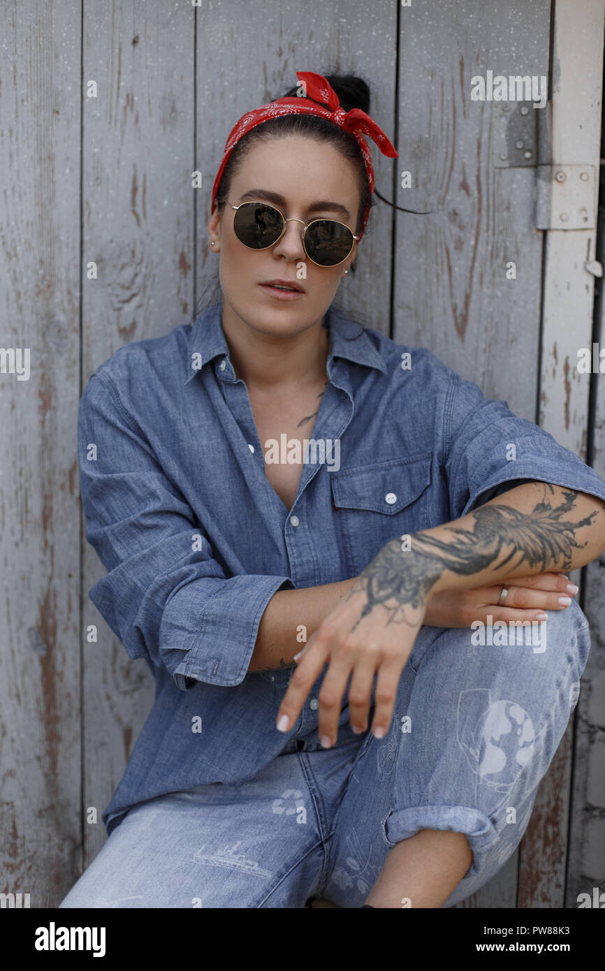 Stilvolle schöne Amerikanische hipster Frau mit einem Tattoo auf der Hand Fashion Denim Shirt und vintage Blue Jeans mit Sonnenbrille sitzt in der Nähe von einem hölzernen Wal Stockfoto
