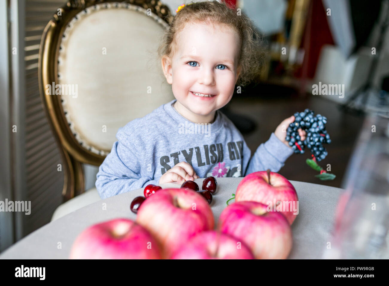 Blue-eyed niedliche Mädchen an einem Tisch sitzen mit Äpfeln, Kirschen, Trauben und lächelnd Stockfoto