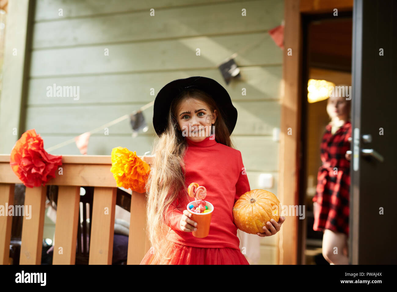 Kleines Mädchen in rot Halloween Kostüme und schwarzen Hut holding Papier Glas mit süßen Leckereien und reife Kürbis Stockfoto