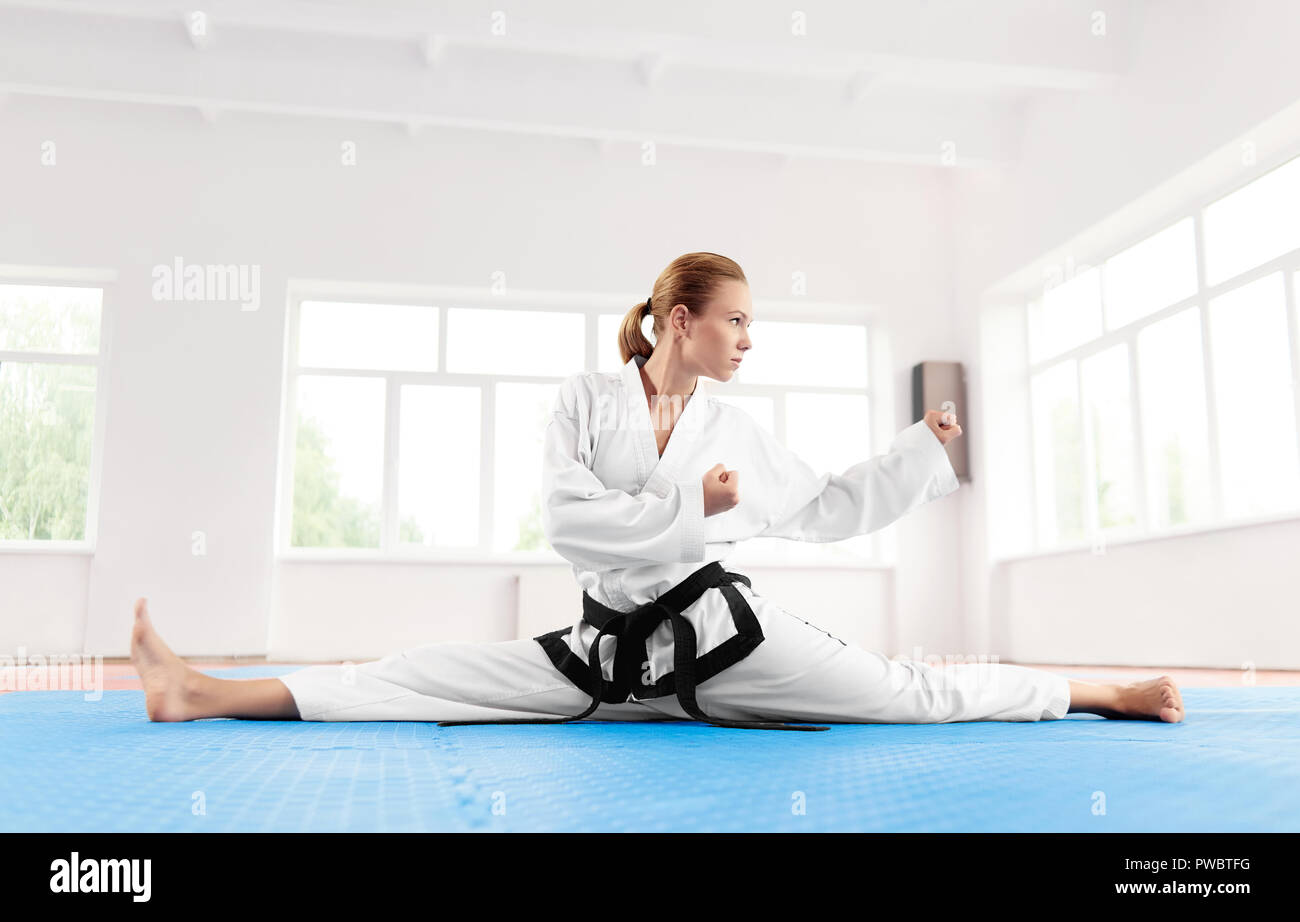 Jungen und Erfolgreichen Karate Frau, tragen im weissen Kimono und schwarzen Riemen dehnen Bein vor hartes Training im Kampf Klasse. Kämpferin sitzen auf Mate und String vor dem Kampf den Wettbewerb. Stockfoto
