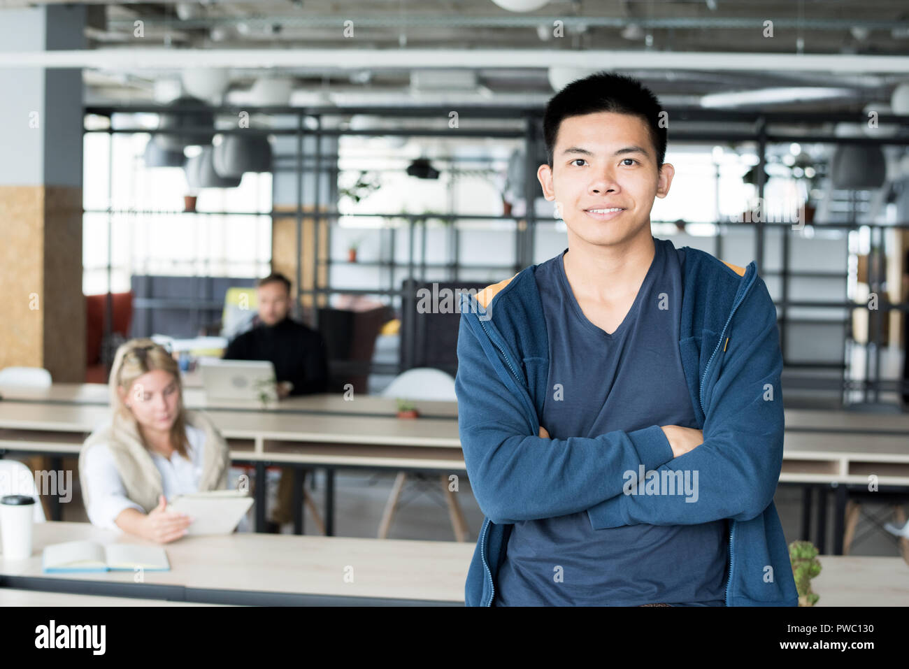 Glückliche junge asiatische männlichen Praktikanten mit verschränkten Armen lehnte sich auf Tabelle und Kamera im Coworking Space Stockfoto