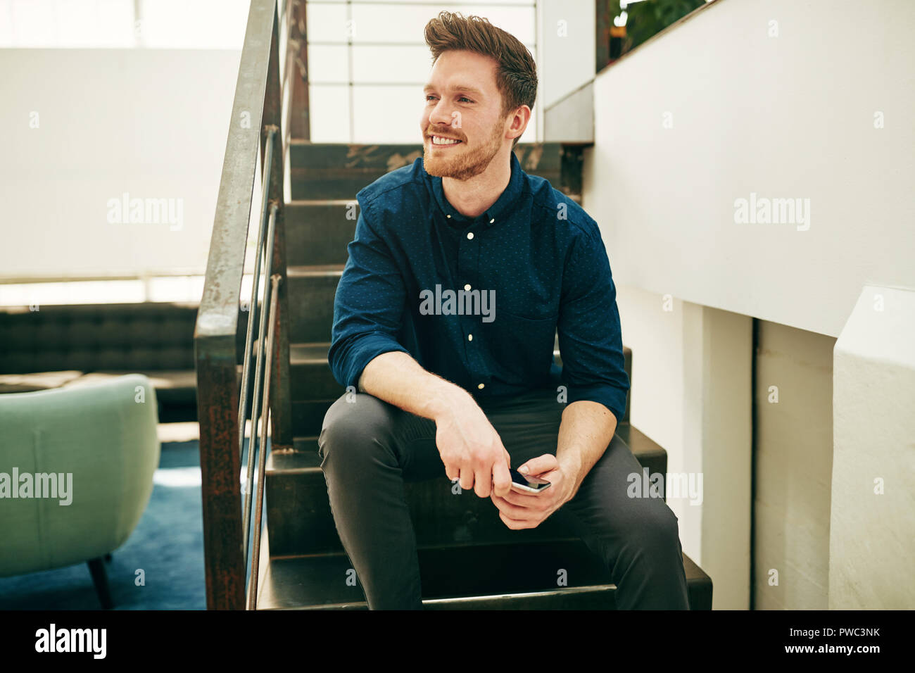 Lächelnde junge Unternehmer mit einem Bart allein sitzen auf der Treppe während seiner Pause in einem modernen Büro mit einem Mobiltelefon Stockfoto