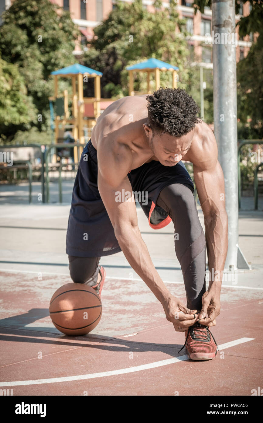 Eine afroamerican junger Mann ohne T-Shirt ist Basketball spielen in einem Park in Madrid im Sommer mittags. Er ist auf seine Knie, seine Schuhe binden mit t Stockfoto