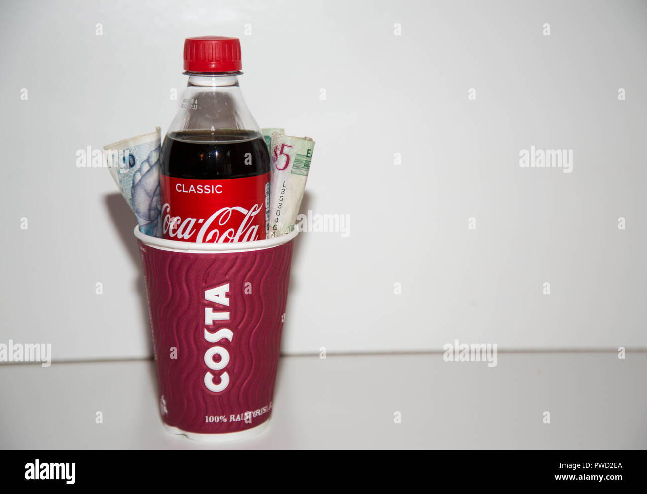 Costa Kaffee Tasse mit Coca Cola Flasche innen mit Bargeld auf jeder Seite auf weißem Hintergrund. Stockfoto