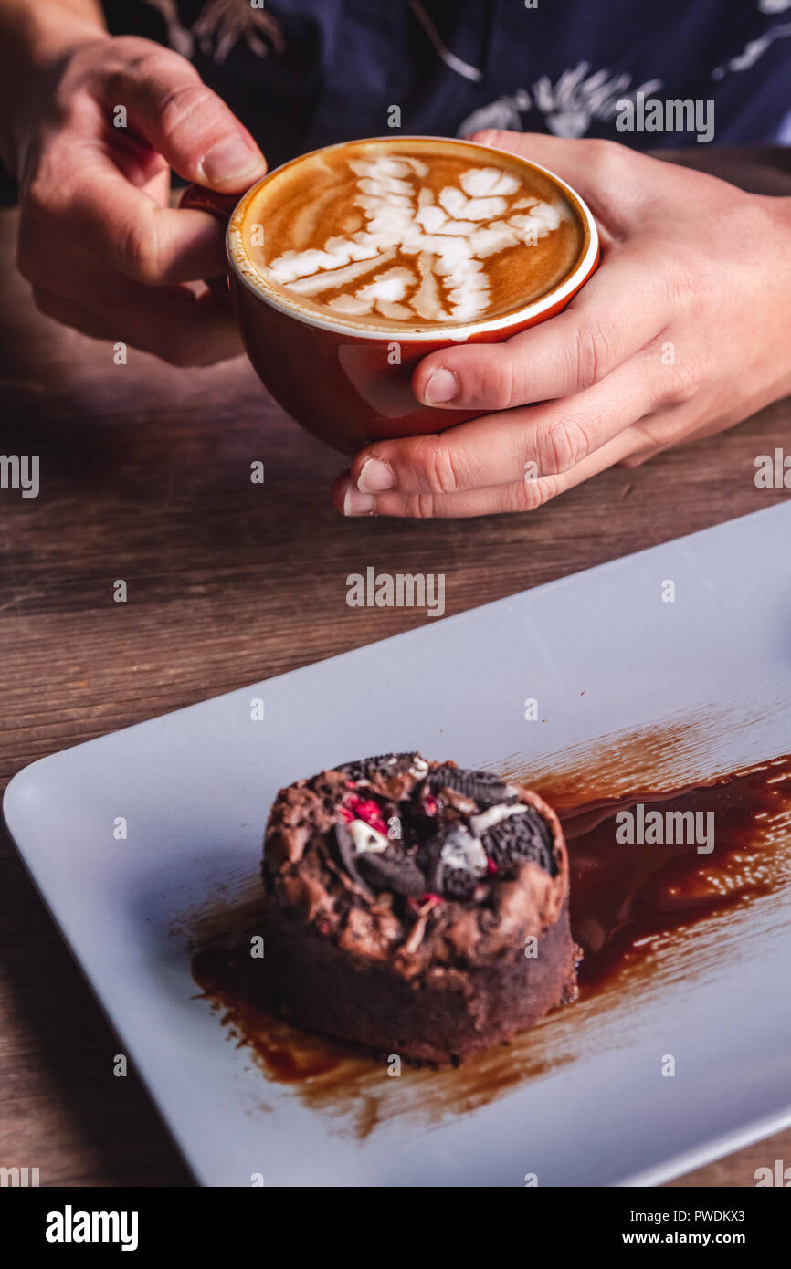 Hände halten Kaffee latte Cup und Schokoladenkuchen auf Tisch Stockfoto