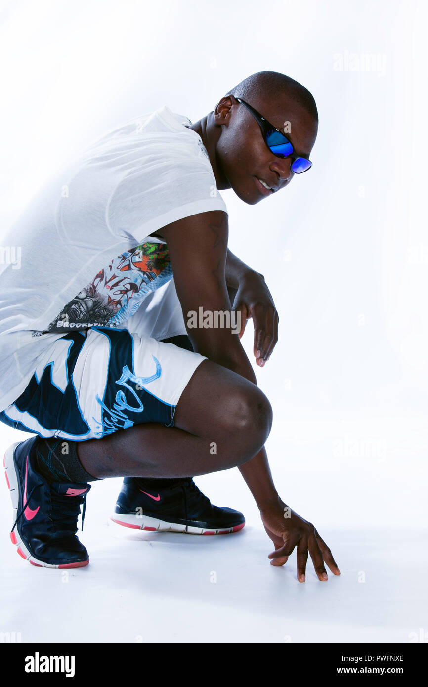 Junge afrikanische Mann in Einem hockenden Position tragen sportliche Kleidung, Turnschuhe und Sonnenbrille in die Kamera schaut Stockfoto