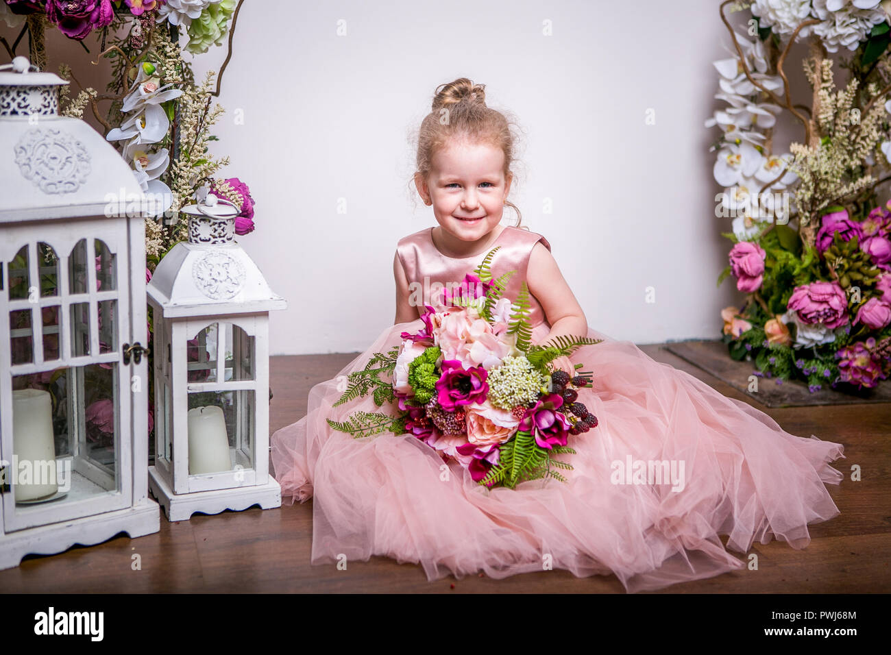 Eine kleine Prinzessin in einem schönen rosa Kleid sitzt auf dem Boden in  der Nähe von Blumen steht und Laternen, hält einen Strauß Pfingstrosen,  Magnolien, Beeren und Grün, und Lächeln Stockfotografie -