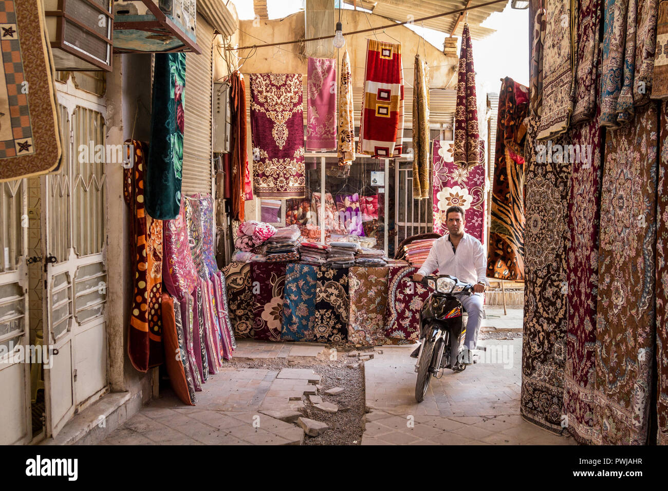 YAZD, IRAN - 18. AUGUST 2015: Mann, der ein Motorrad durch Geschäfte verkaufen Teppiche & Teppiche in Yazd Khan Basar umgeben. Yazd ist eine der wichtigsten Städte Stockfoto