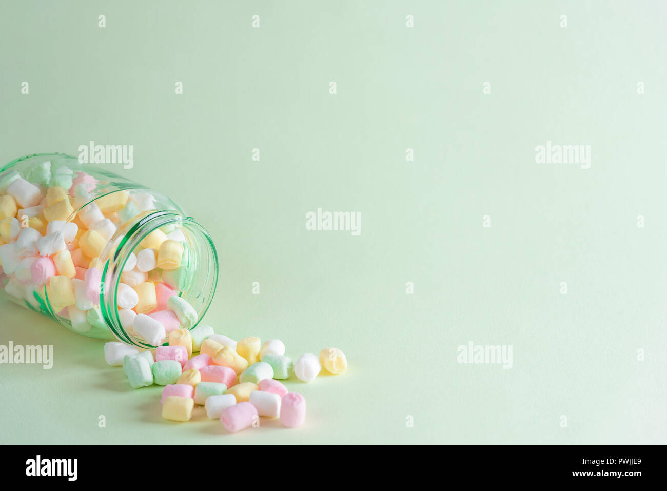 Winter behandelt minimal style Bild mit einem Glas voll in verschiedenen Farben kleine Marshmallows über eine grünliche Papier Hintergrund verschüttet. Stockfoto