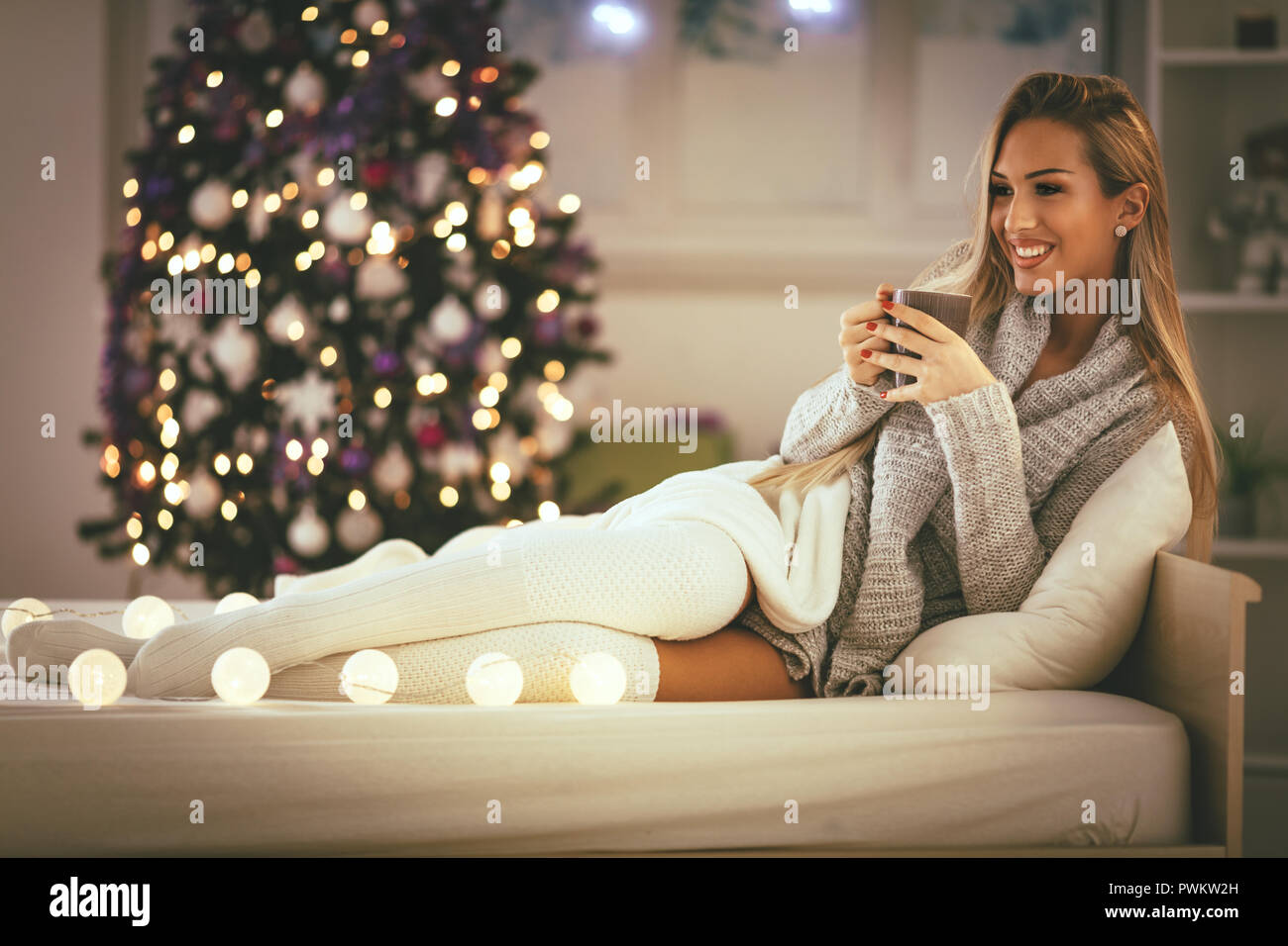 Süße junge lächelnde Frau mit Tasse Kaffee und Sitzen im Bett mit Weihnachten bubble Lights. Stockfoto