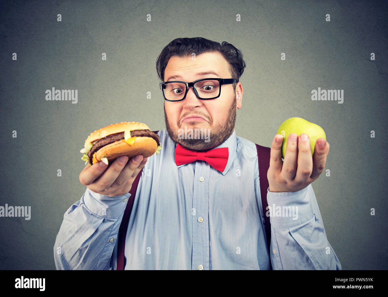 Junge mollig Mann in Bow Tie und Gläser mit Apfel und Burger mit Wahl verwirrt auf der Suche Stockfoto