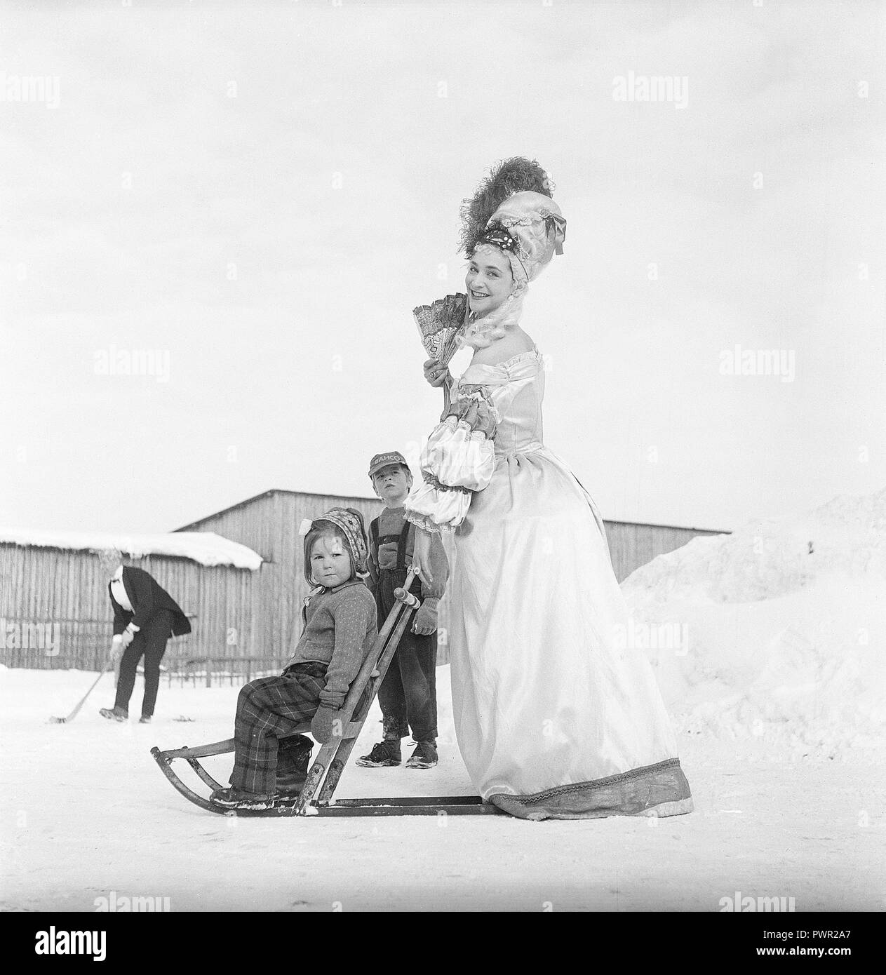 Winter in den 50er Jahren. Eine seltsame Szene mit einer Frau wie auf einem Filmset, in historischem Kostüm. Sie steht auf einem kicksled oder Funken mit einem kleinen Mädchen in den vorderen Sitzen. Schweden 1950. Foto Kristoffersson ref BS 22-9 Stockfoto